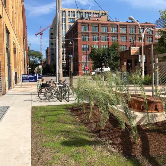 Sidewalk path leading north in front of CUPPA Hall with newly planted grasses to the east, the building to the west, and bike parking and the building sign ahead.