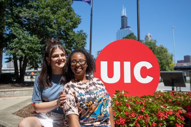 Two UIC students posing in front of the UIC logo red dot sign and the Willis Tower in the background.