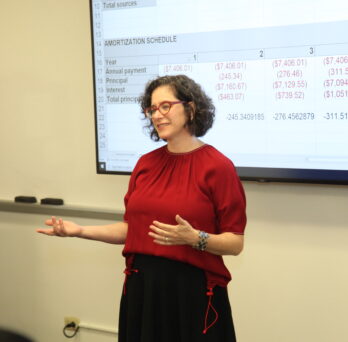 Urban planning and policy professor, Rachel Weber, in red shirt in front of a screen with a development finance spreadsheet in the classroom
                  