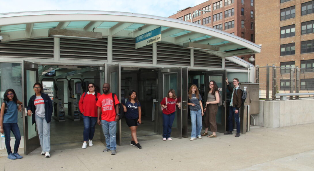 Nine CUPPA students in various UIC teeshirts and sweatshirts stand in front of the Entry of the CTA station on the Peoria Street bridge