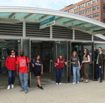 Nine CUPPA students in various UIC teeshirts and sweatshirts stand in front of the Entry of the CTA station on the Peoria Street bridge
                  