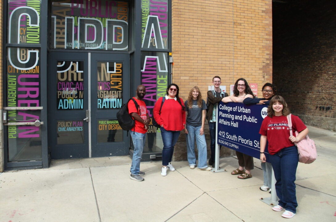 Seven CUPPA students pose outside of CUPPA Hall near the official university sign for the building entry.