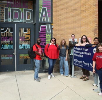 Seven CUPPA students pose outside of CUPPA Hall near the official university sign for the building entry. 