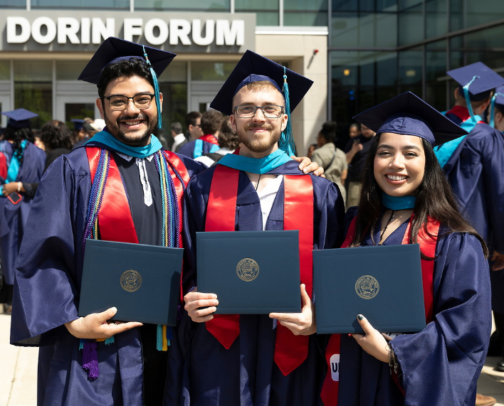 Three graduates in navy blue caps and gowns with red UIC stoles stand in front of the UIC Dorin Forum.