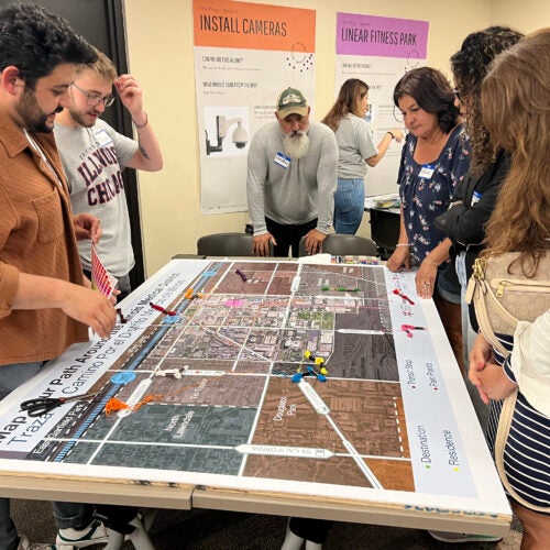 Student in an orange shirt at a table displaying design maps of a neighborhood with community members at the table as well.