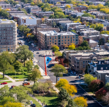 Photo of west side Chicago neighborhood with trees, green grass, and both tall and medium size brick buildings.
                  