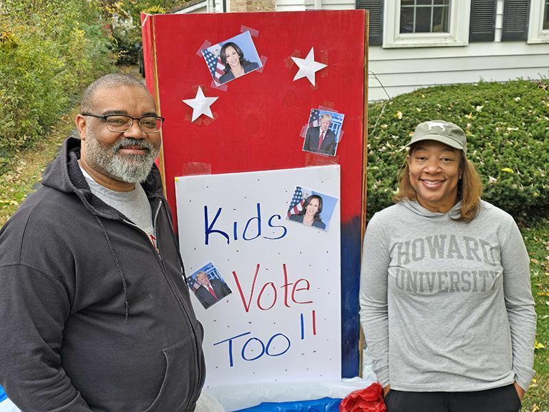 Joe Hoereth stands to the left of a red sign that says, Kids Vote Too. Joe's wife stands to the right of the sign in a gray Howard University sweatshirt.