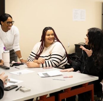 the course co-instructor in a while sweater stands at a table with three different students 