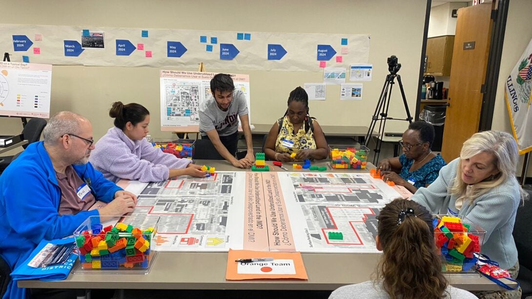 Seven people are gathered around a table with maps and drawings. Two students appear to be explaining the maps.