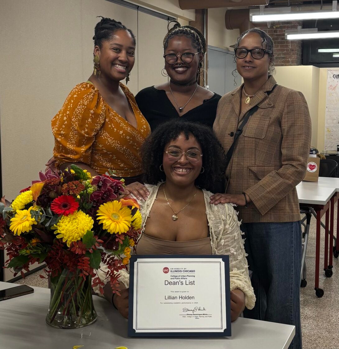 Student seated with her certificate of achievement and a bouquet of flowers on her left with her three guests standing behind her.