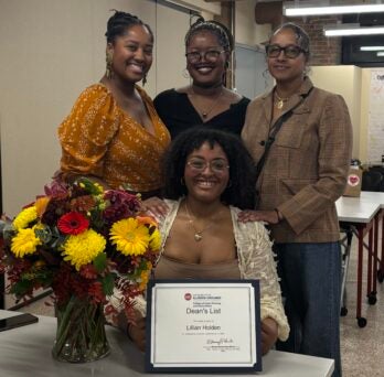 Student seated with her certificate of achievement and a bouquet of flowers on her left with her three guests standing behind her.
                  